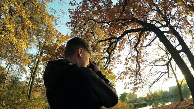 Young caucasian guy boy teen photographer takes pictures in the sunny autumn park among the trees 