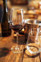 Close up of a still life of an elegant long-stemmed goblet with red wine and a glass of water on a textured wooden table. An ice cooler and a dark glass bottle in the background. Romantic atmosphere.