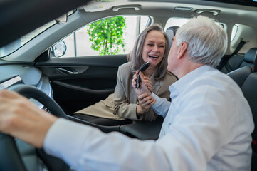 An elderly man holds the keys while sitting in a new car. A gray-haired married couple is happy about buying a car.