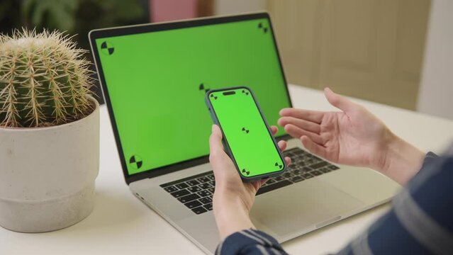 Hands holding a smartphone with a green screen in front of a laptop with a matching green screen, beside a potted cactus on a desk