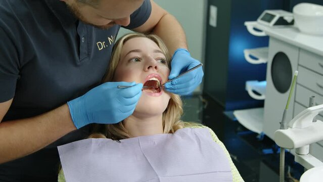 Young dentist in safety gloves using tooth scraper, concave mirror, checking condition of teeth of woman sitting in armchair with patient napkin on chest. High quality 4k footage