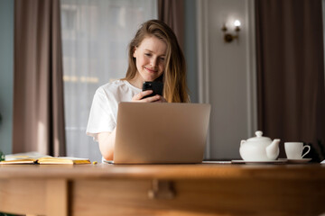 Smiling young woman using smartphone behind laptop, with a teapot and cup, in a warm home setting.