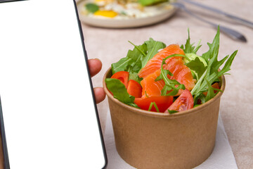 Salmon salad with tomatoes and arugula in cardboard box and smartphone with blank screen