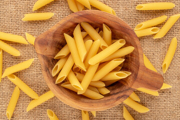 Raw pasta, penne, in a wooden cup on a jute cloth, macro, top view.