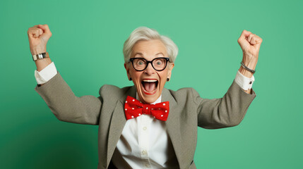 Person with short white hair and glasses, wearing a red bow tie and a mint jacket, celebrating enthusiastically with fists pumped and a wide-open mouth, against a vibrant green background. - obrazy, fototapety, plakaty