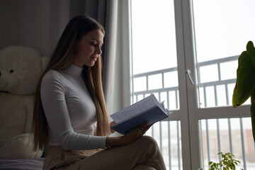 Young woman sitting on modern sofa in front of window relaxing in her living room reading book