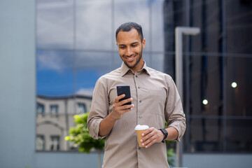 Businessman walking on break outside office building, male holding phone in hands, using app on smartphone to browse social network, holding cup of hot coffee drink in hand, smiling joyfully.