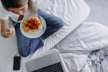 Top view of Asian Thai woman eating homemade pasta and sitting on sofa bed in room apartment while watching series movie by laptop, using notebook working alone in day time. - obrazy, fototapety, plakaty