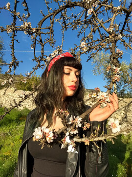 A sultry pin-up dons a black dress with red accents, posing gracefully beside a blossoming almond tree branch.