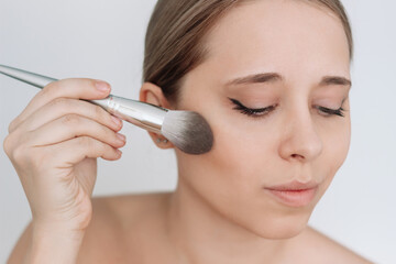 Close up of a young caucasian blonde woman using a brush applying blush to the skin isolated on a white background. A girl doing makeup. Cosmetic beauty concept