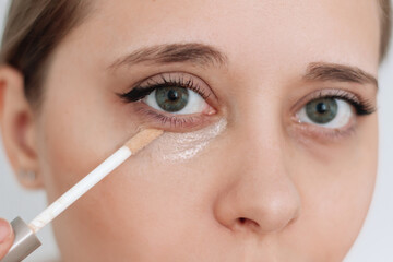 Close-up of a young caucasian attractive woman applying the concealer for dark eyes with the brush. Bruises under the eyes. Foundation cream hiding spot imperfections on female face
