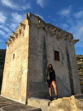 Vintage pin-up, donned in black and red, poses by a medieval tower on Gargano with a bold slit and captivating smile, enhancing the allure amid the historic setting.