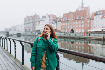 Young happy redhead woman making phone call over mobile phone in the city. Middle age woman talking on smartphone on urban background. Traveling Europe in autumn