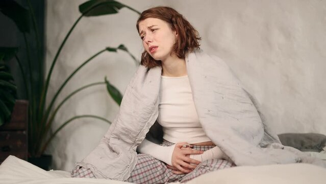 Sick unhealthy young woman student clasping belly and suffering from severe abdominal pain periods spasm irritable bowel syndrome of diet sitting on bed at light home