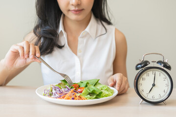 Intermittent fasting with clock, health asian young woman, girl weight loss, eating green fresh vegetable salad on dish, plate with eat healthy of breakfast food in morning, lunch on a table at home.