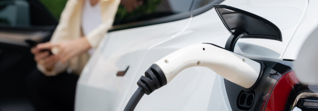 Young Woman Use Smartphone To Pay For Electricity At Public EV Car Charging Station Green City Park. Modern Environmental And Sustainable Urban Lifestyle With EV Vehicle. Panorama Expedient