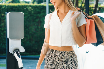 Young woman travel with EV electric car charging in green sustainable city outdoor garden in...