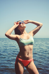 A happy tourist in a swimsuit and sunglasses likes to take pictures in nature as a souvenir. A female traveler poses on the beach by the sea and enjoys the sun.