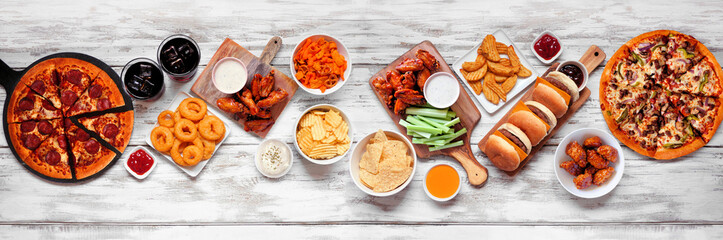 Junk food table scene. Pizza, hamburgers, chicken wings and salty snacks. Top view over a white wood banner background.