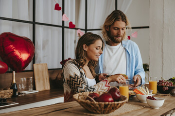 Beautiful young couple having breakfast at the kitchen with red heart shape balloons on background