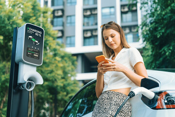 Young woman travel with EV electric car charging in green sustainable city outdoor garden in...