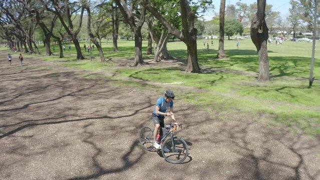 Man in cycling helmet and sportswear standing with bicycle on unpaved road in the park and using mobile phone while having a break during ride. Aerial shot made from drone
