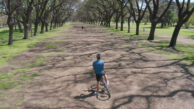 Man standing with bicycle on dirt road in the park, using map on phone and looking around while planning route for ride. High angle shot from flying drone