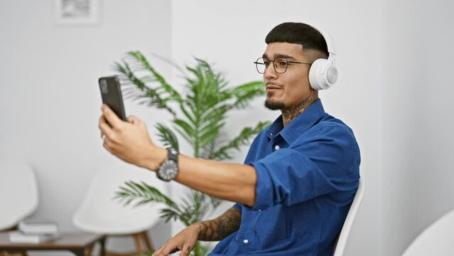 Handsome young latin man immersed in passionate business conversation during video call, sitting in the waiting room, his tattoo peeking from under the sleeve, focused on his phone screen.