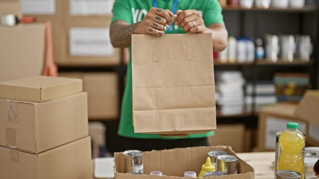 Tattooed hispanic adult man volunteers at local charity center, holding a paper bag of canned food donations, exemplifying community unity and altruism