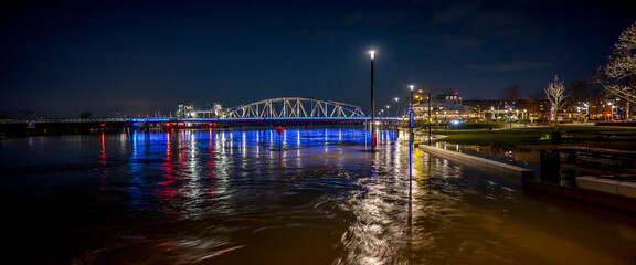 Wide panorama of colourful decoration on steel draw bridge in the background with high water levels of river IJssel flooded countenance boulevard of tower town Zutphen at nighttime with street lights