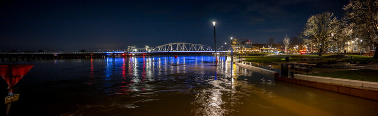 Fototapeta na wymiar Wide panorama of colourful decoration on steel draw bridge in the background with high water levels of river IJssel flooded countenance boulevard of tower town Zutphen at nighttime with street lights