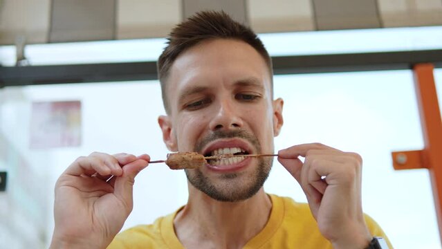 Hispanic man bites from wooden skewer and chews grilled kebab. Portrait of handsome man with stubble having lunch on terrace in cafe, up view. Eating of main dish of minced meat with seasonings.