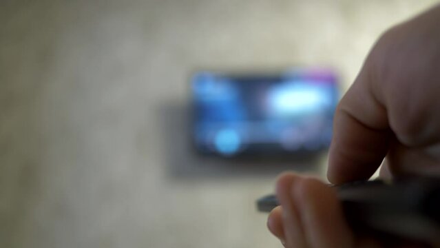 Close-up of a male hand with a TV remote control switches TV channels and turns off the TV. A man is resting on the bed and watching TV.