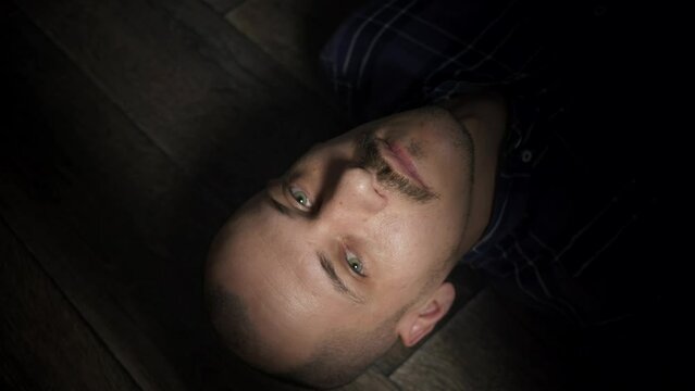 A bald and mustachioed man lies down on the floor and looks up. A tired, skinny man looks at the camera while lying on the floor. The emotionless look of a person.