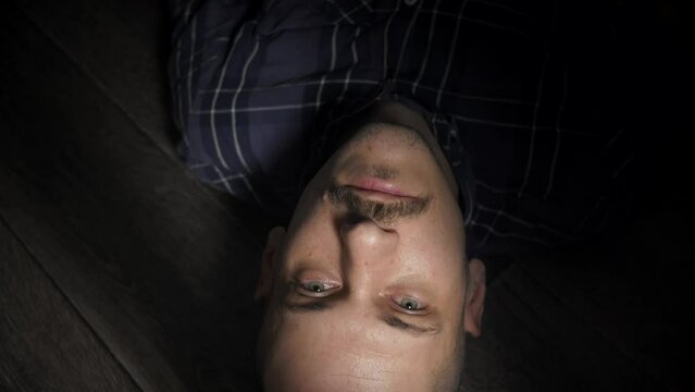 A bald and mustachioed man opens his eyes and looks at the camera while lying on the floor. A tired, skinny man looks up while resting on a wooden floor. Emotionless look of a man.