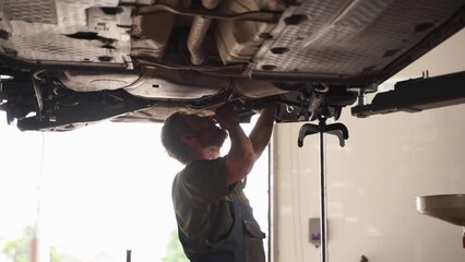 Auto mechanic works on car suspension in repair shop. Fixes vehicle underside, inspects springs, shock absorbers. Professional garage service, maintenance. Car lifted, technician examines chassis.