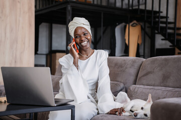 Smiling African woman in a headscarf having a pleasant conversation on a mobile phone stroking dog...