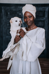 Elegant woman in white with a headscarf holding a small white dog, African girl in turban carrying pet feels affection.