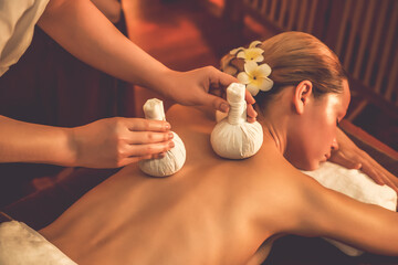 Hot herbal ball spa massage body treatment, masseur gently compresses herb bag on woman body. Tranquil and serenity of aromatherapy recreation in warm lighting of candles at spa salon. Quiescent - obrazy, fototapety, plakaty