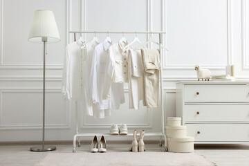 Rack with different stylish women`s clothes, shoes, lamp and dresser near white wall in room