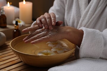 Obraz na płótnie Canvas Woman applying scrub onto her hands in spa, closeup. Bowl of water and flowers on wooden table