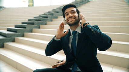Project manager listening and enjoy music from headphone while sitting at stairs. Professional businessman wearing suit outfit while dancing and moving to music. Happy man listen funny song. Exultant. - obrazy, fototapety, plakaty