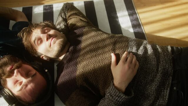 Two young men lying on the floor, wearing headphones, listening to music with eyes closed. Concept of interracial friendship or male homosexual couple