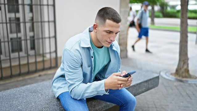 Young hispanic man using smartphone while sitting on bench in urban city setting.