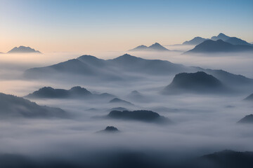 A mystic view of a mountain valley filled with early morning fog, with the tops of mountains peering through and the first light of dawn casting a soft glow.