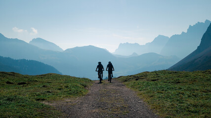 Silhouettes of two men mountainbiking in the Karwendel mountains in front of blue mountain layers during sunny blue sky day in summer, Tyrol Austria. - 704096896