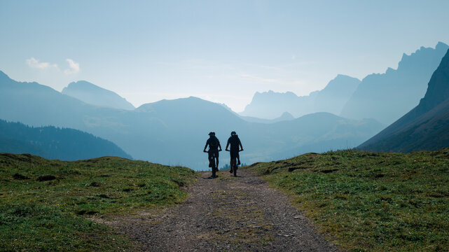 Silhouettes of two men mountainbiking in the Karwendel mountains in front of blue mountain layers during sunny blue sky day in summer, Tyrol Austria.