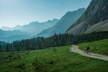 Mountainbiking along gravel road in the Karwendel mountains in front of blue mountain layers during sunny blue sky day in summer, Tyrol Austria. - 704097602