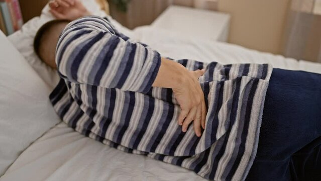 A young hispanic man in a striped shirt lying in bed at home holding his back, possibly indicating pain or discomfort.