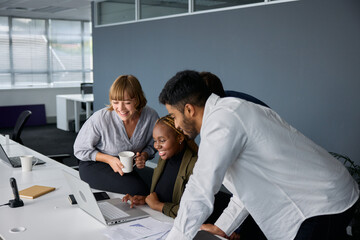 Four young adults in businesswear smiling while working on laptop at desk in office